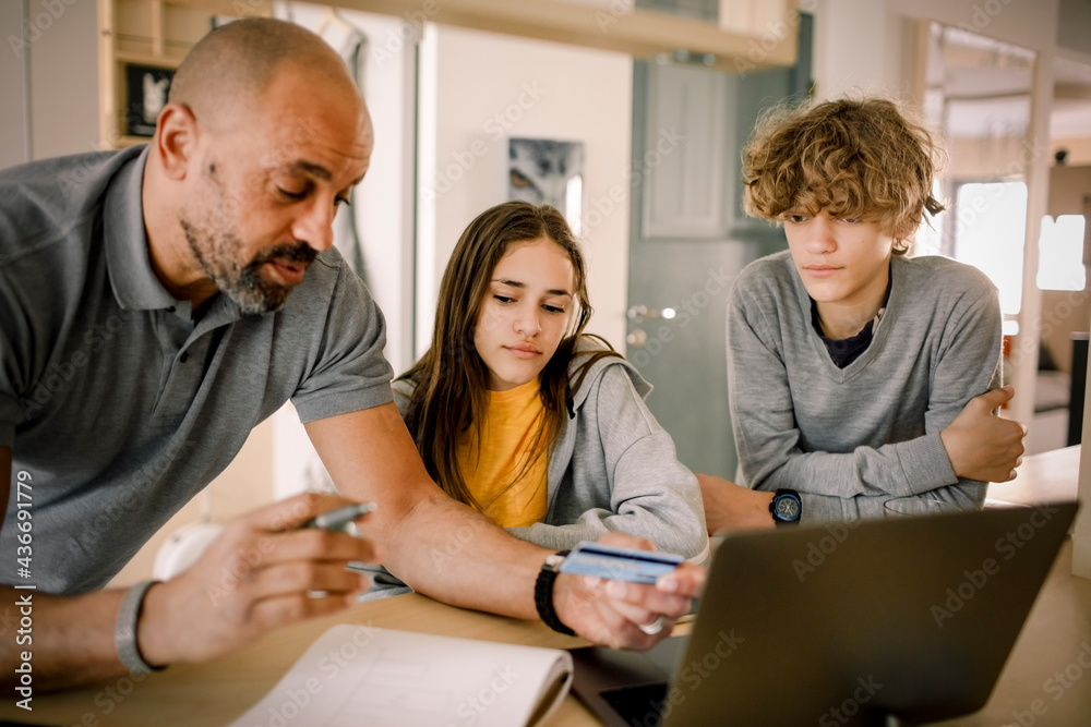 Brother and sister looking at father explaining finance with credit card at kitchen island Brother and sister looking at father explaining finance with credit card at kitchen island