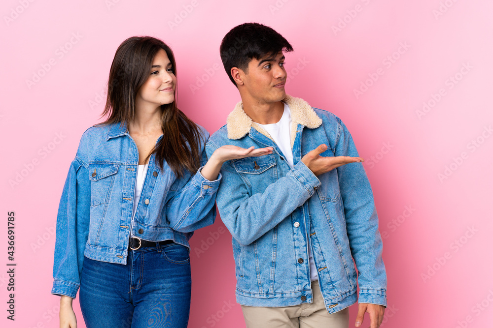 Young couple over isolated pink background presenting an idea while looking smiling towards Young couple over isolated pink background presenting an idea while looking smiling towards