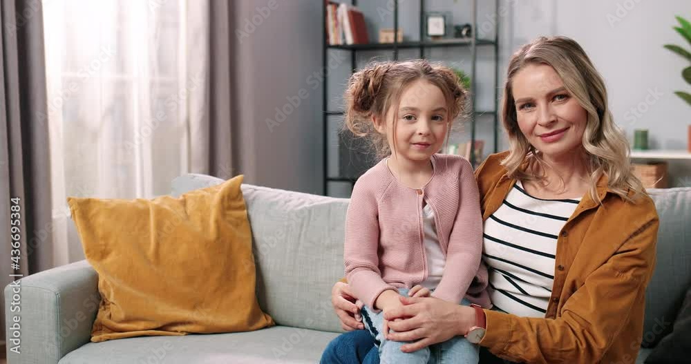 Portrait of joyful lovely Caucasian little adorable girl sitting on couch in living room with pretty mother looking at camera and smiling. Caring mom with kid spending time together, family concept Portrait of joyful lovely Caucasian little adorable girl sitting on couch in living room with pretty mother looking at camera and smiling. Caring mom with kid spending time together, family concept