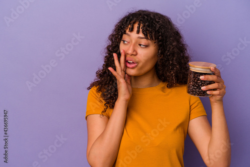 Young mixed race woman holding a coffee beans bottle isolated on purple background is saying a secret hot braking news and looking aside