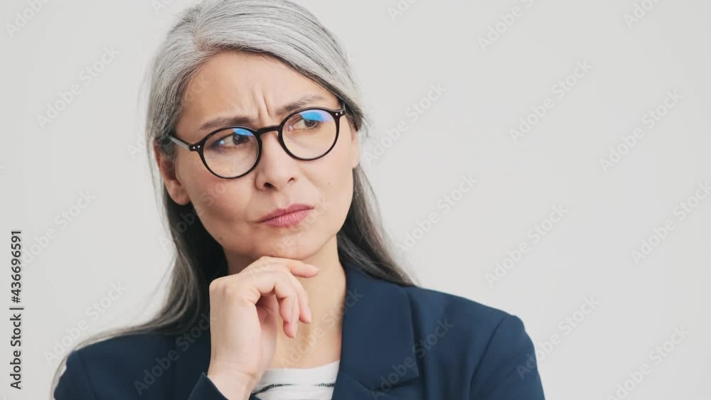A puzzled elder businesswoman is shrugging shoulders standing isolated over grey wall in the studio