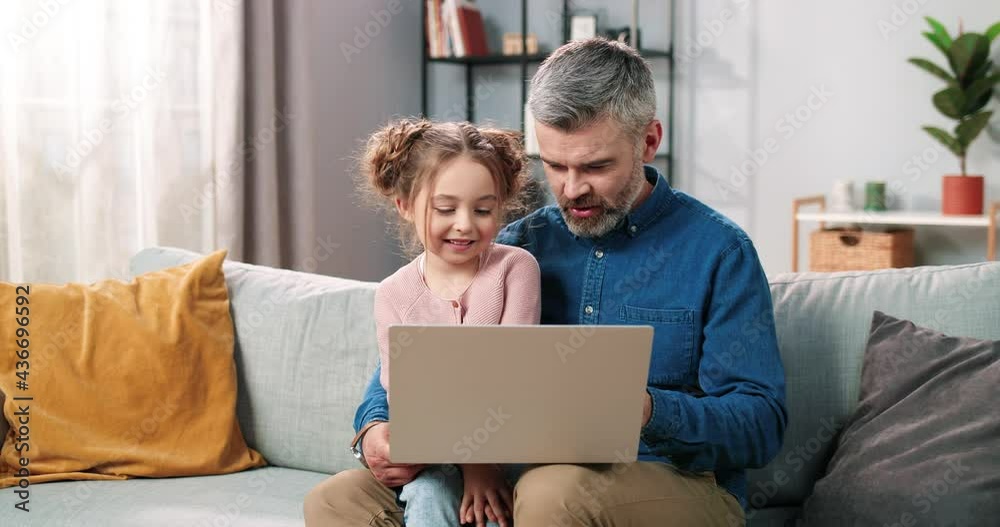 Portrait of happy nice Caucasian little daughter and caring father spending time together at home using laptop online. Dad and cute small kid girl tapping on computer browsing on internet Portrait of happy nice Caucasian little daughter and caring father spending time together at home using laptop online. Dad and cute small kid girl tapping on computer browsing on internet