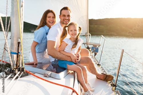 Cheerful Family Of Three Relaxing Embracing Sitting On Yacht Deck © Prostock-studio