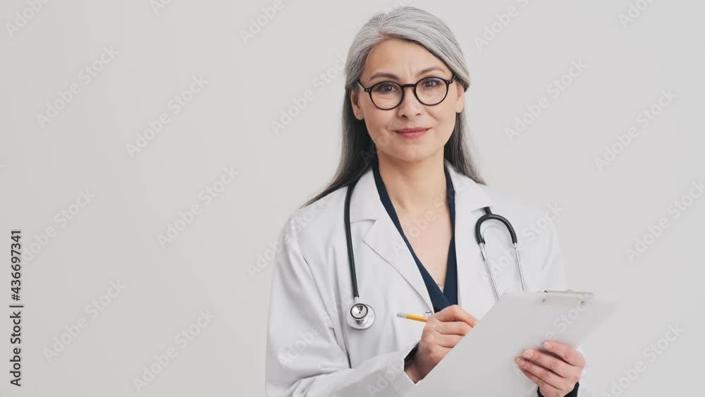 A pleased senior woman doctor is holding a folder with documents standing isolated over grey wall in the studio