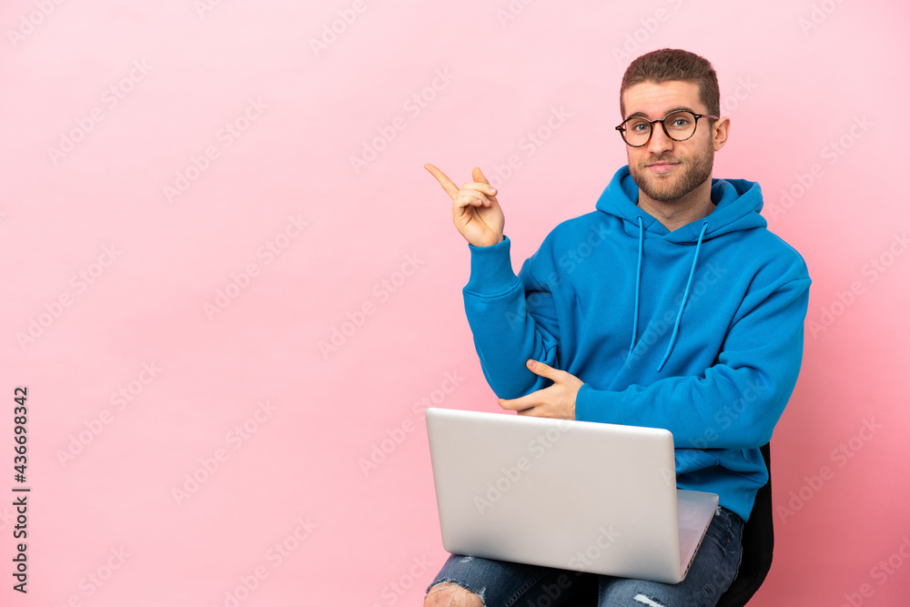 Young man sitting on a chair with laptop pointing finger to the side