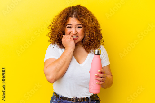 Young latin woman holding a thermos isolated on yellow background biting fingernails, nervous and very anxious.