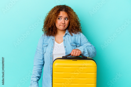 Young latin traveler curvy woman holding a suitcase isolated on blue background confused, feels doubtful and unsure. photo