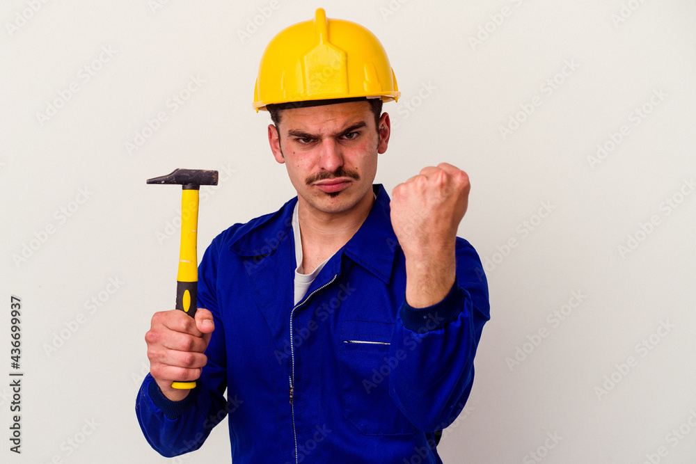 Young caucasian worker man holding a hammer isolated on white background showing fist to camera, aggressive facial expression. Young caucasian worker man holding a hammer isolated on white background showing fist to camera, aggressive facial expression.