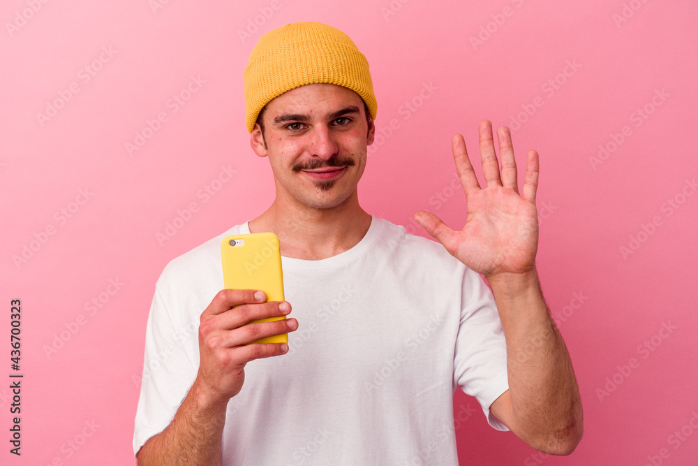 Young caucasian man holding a mobile phone isolated on pink background smiling cheerful showing number five with fingers. Young caucasian man holding a mobile phone isolated on pink background smiling cheerful showing number five with fingers.