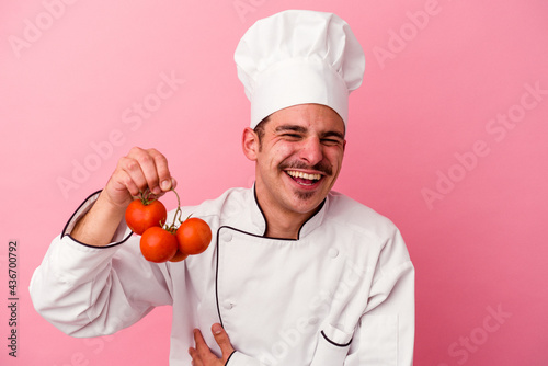 Young caucasian chef man holding tomatoes isolated on pink background laughing and having fun.