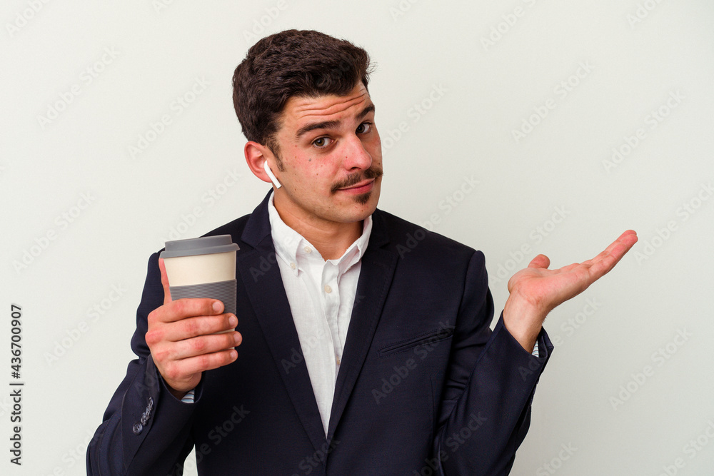 Young business caucasian man wearing wireless headphones and holding take way coffee isolated on white background showing a copy space on a palm and holding another hand on waist.