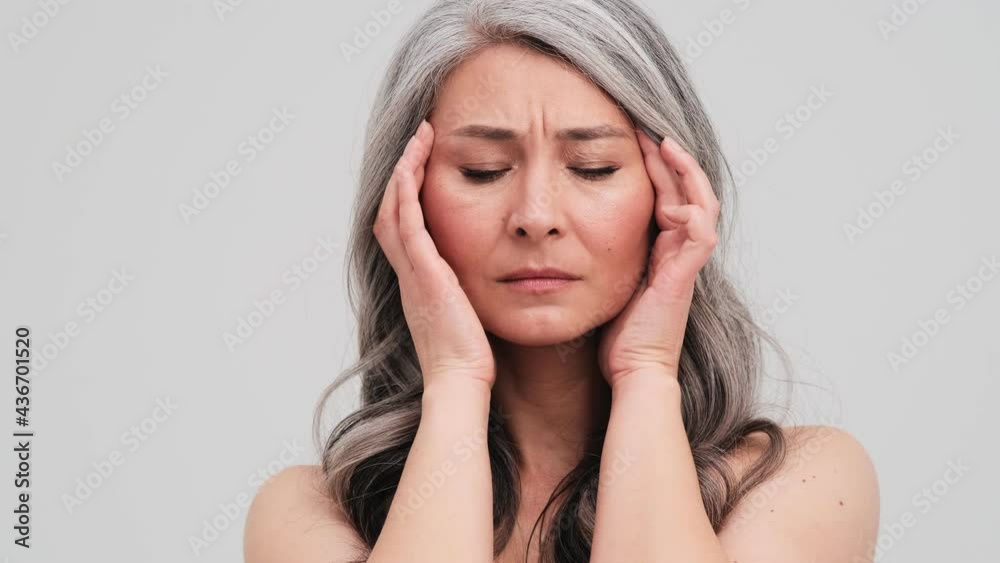 A serious elder woman is massaging her temples standing isolated over grey wall in the studio