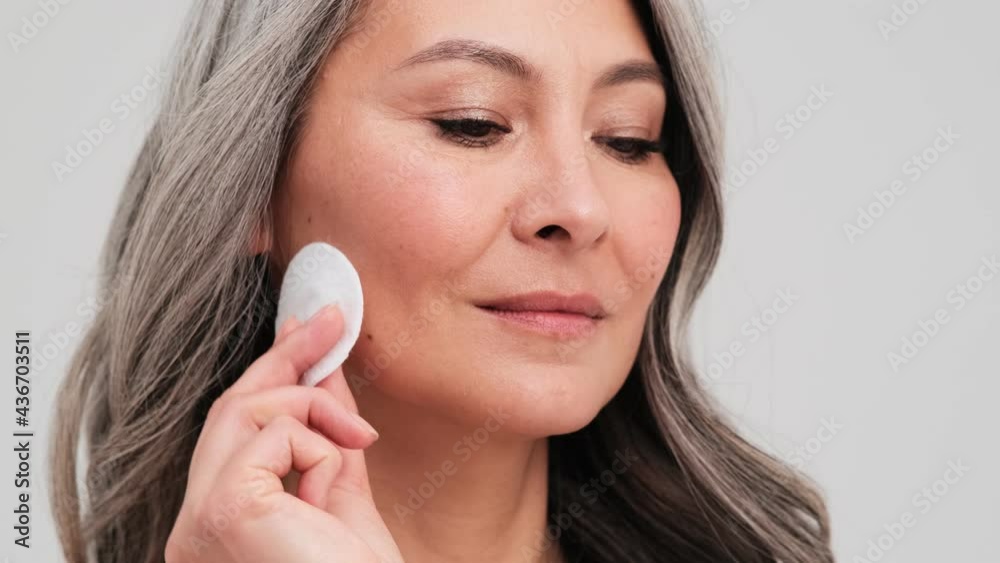A close-up view of a pleased elder woman is using a face sponge standing isolated over grey wall in the studio A close-up view of a pleased elder woman is using a face sponge standing isolated over grey wall in the studio