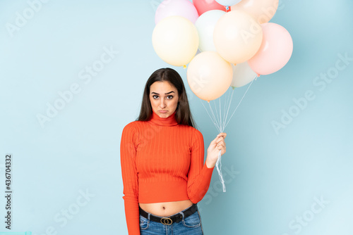 Young woman catching many balloons isolated on blue background sad © luismolinero