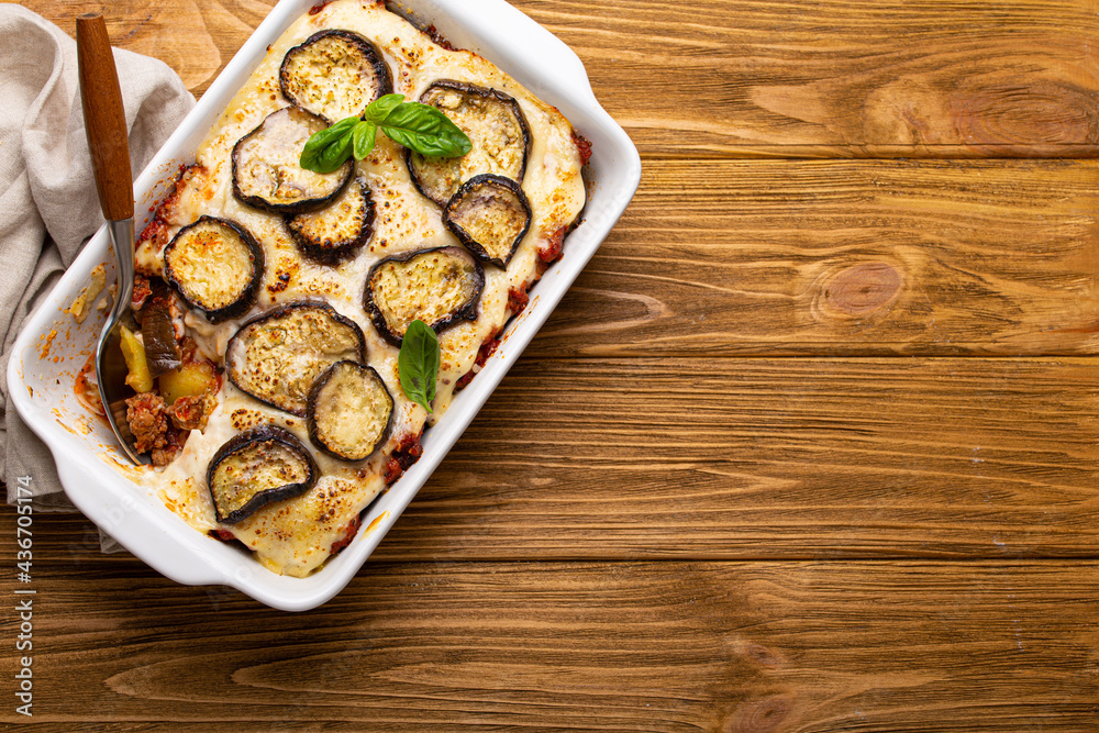 Greek mediterranean dish Moussaka with baked eggplants, ground beef in white ceramic casserole with napkin on wooden rustic background from above, traditional dish of Greece