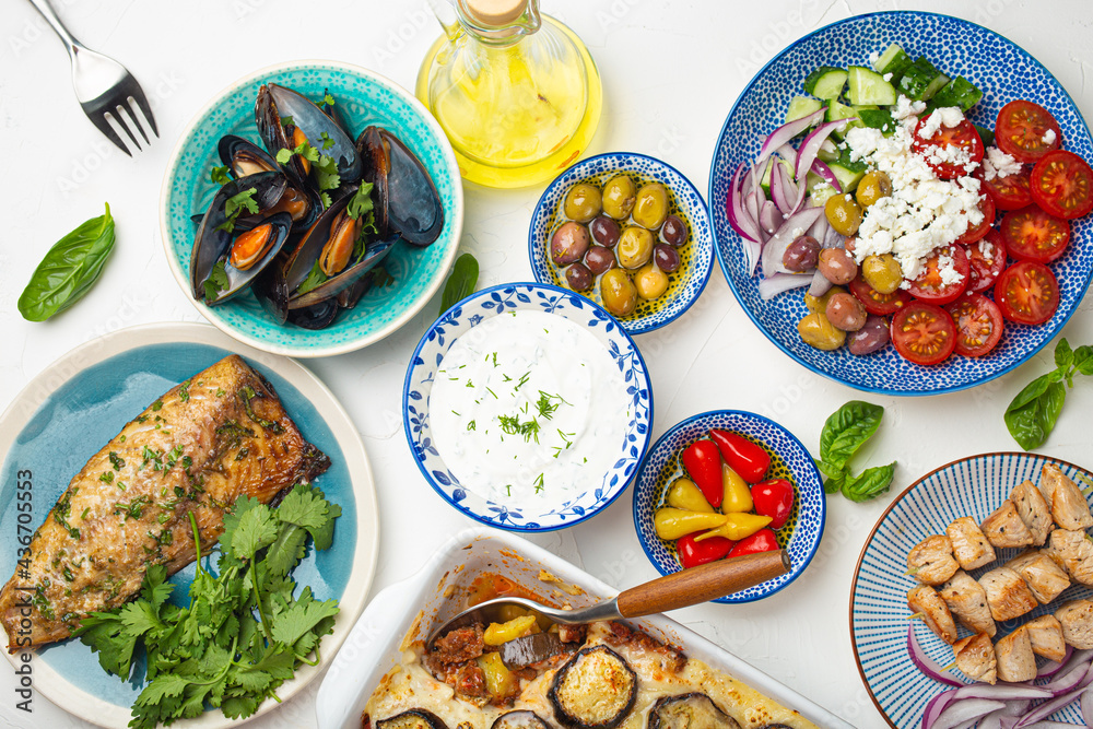 Assorted Greek dishes on white stone background from above, moussaka, grilled fish, souvlaki, greek salad, steamed mussels with herbs, appetizers of Greece from above 