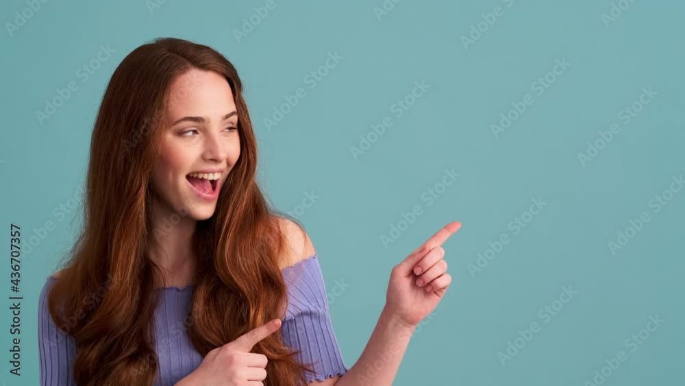 A smiling woman is pointing with fingers to the side standing isolated over blue wall in the studio A smiling woman is pointing with fingers to the side standing isolated over blue wall in the studio