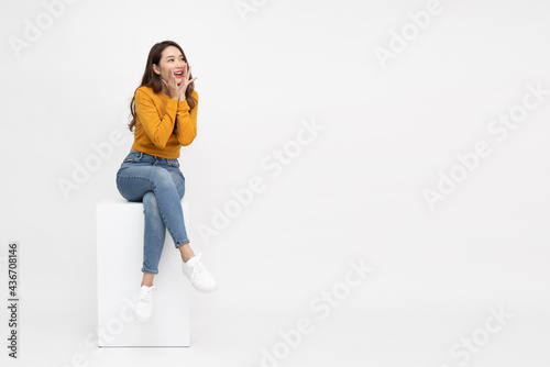Fotografie Portrait of excited screaming young asian woman sitting on white box isolated ov