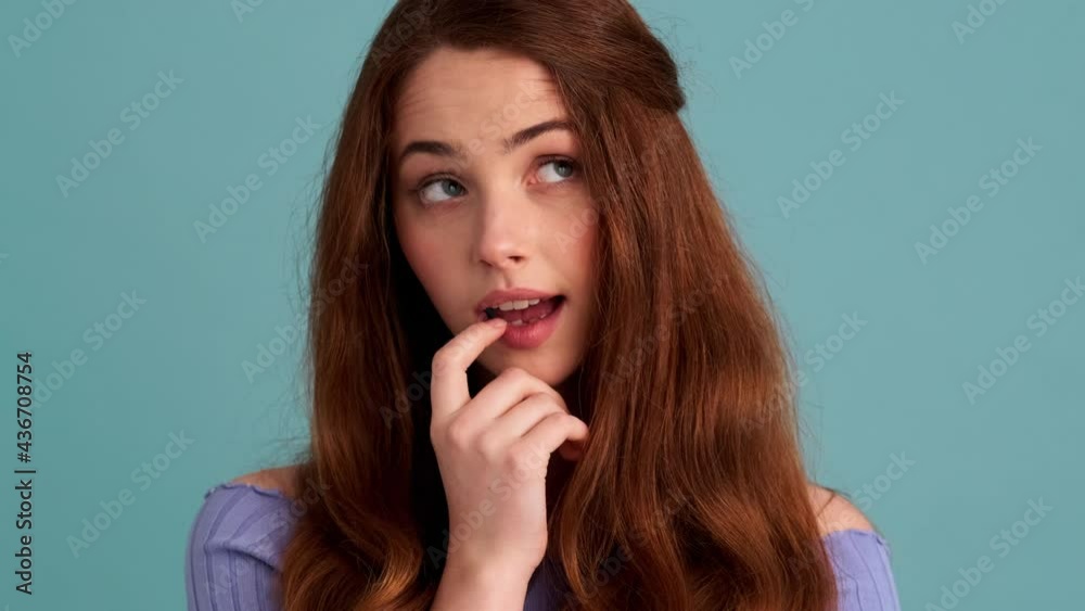 A dreaming woman is looking to the camera standing isolated over blue wall in the studio A dreaming woman is looking to the camera standing isolated over blue wall in the studio