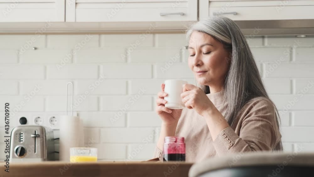 A pleased elder woman is drinking a cup of tea sitting in the kitchen at home A pleased elder woman is drinking a cup of tea sitting in the kitchen at home