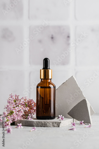 Glass brown serum bottle with a pipette for skin care with branches and flowers of lilac on a gray background. Health and beauty concept