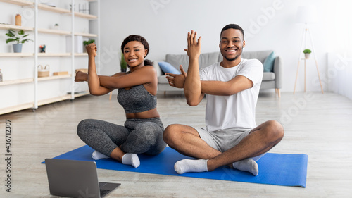 Online yoga class. Young black couple stretching arms, sitting in lotus pose, training together at home, using laptop