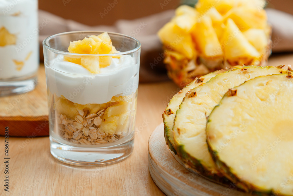 Delicious pineapple dessert. Breakfast dessert with oat granola, greek yogurt and pineapple in layers in glass with ingredients chopped fresh juicy pineapple on wooden table. Delicious pineapple dessert. Breakfast dessert with oat granola, greek yogurt and pineapple in layers in glass with ingredients chopped fresh juicy pineapple on wooden table.