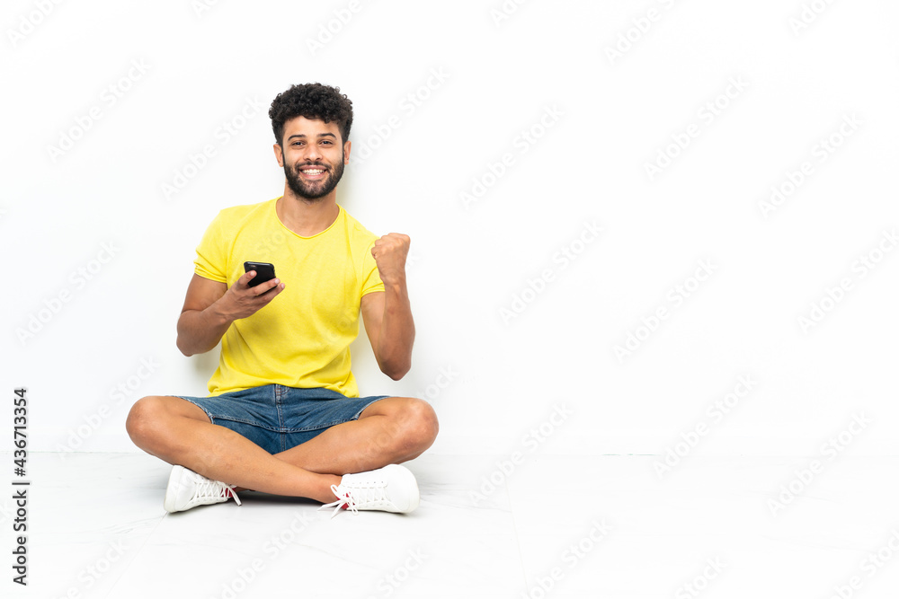 Fototapeta premium Young Moroccan handsome man sitting on the floor over isolated background with phone in victory position