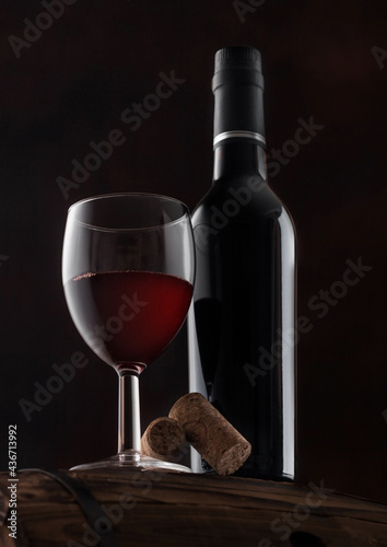 Bottle and glass of red wine with corks and vintage corkscrew on top of wooden barrel on black.