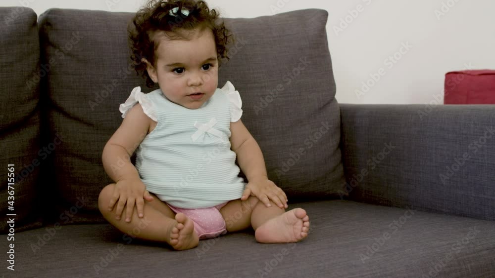 Cute cheerful infant sitting on sofa, trying to dance. Caucasian curly-haired girl having fun with herself at home. Toddler, having fun concept. 