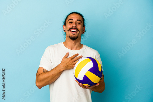Young caucasian man with long hair isolated on blue background laughs out loudly keeping hand on chest.