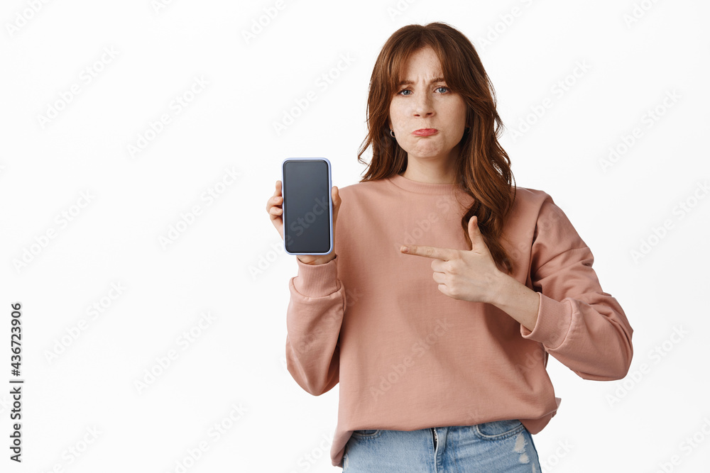 Angry and disappointed girl pointing finger at smartphone screen, complaining on something posted online, bad app or photo, standing upset against white background