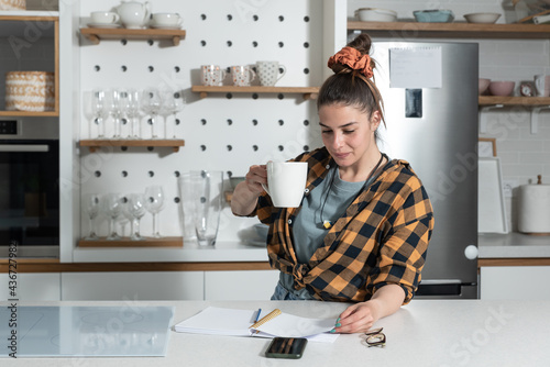 Young bride woman before marriage stands in the kitchen drinking coffee and makes a list of friends invitations for her bachelorette party before the wedding photo