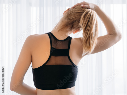 Fitness therapy. Home sport. Morning exercise. Active lifestyle. Back view of athletic woman in sportswear black mesh top stretching neck on light white window background.