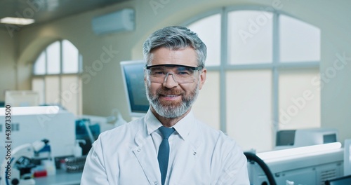 Close up portrait of Caucasian bearded middle-aged male doctor standing in clinic laboratory looking at camera and smiling. Handsome happy man lab specialist at workplace in hospital. Medic concept