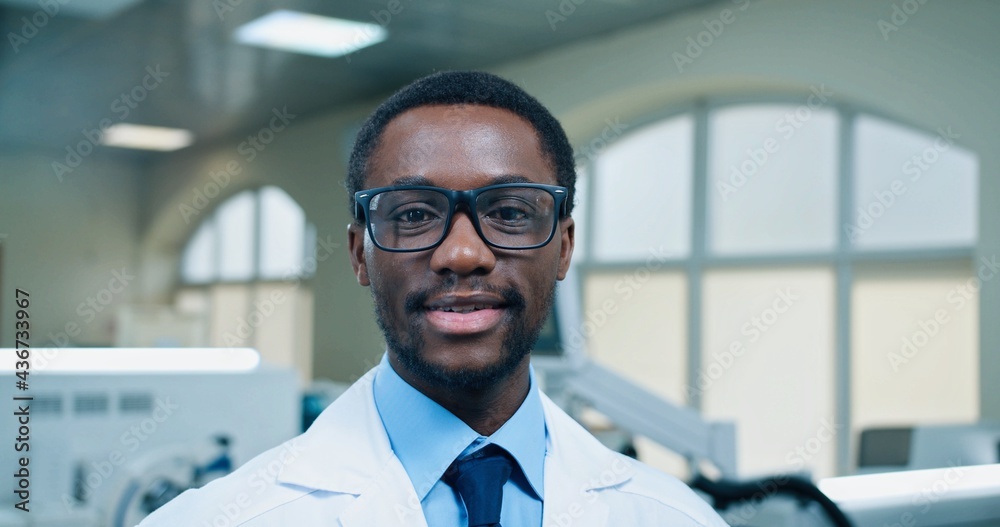 Close up of African American joyful young handsome male doctor in glasses looking at camera and smiling at workplace in hospital lab. Medical worker, laboratory specialist, medicine concept