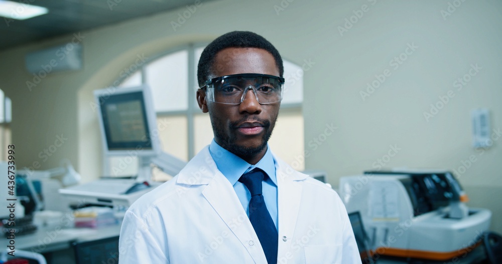 Fototapeta premium Portrait of happy African American young handsome male physician in goggles looking at camera and smiling standing at work in hospital lab. Medical profession, laboratory specialist, medicine concept