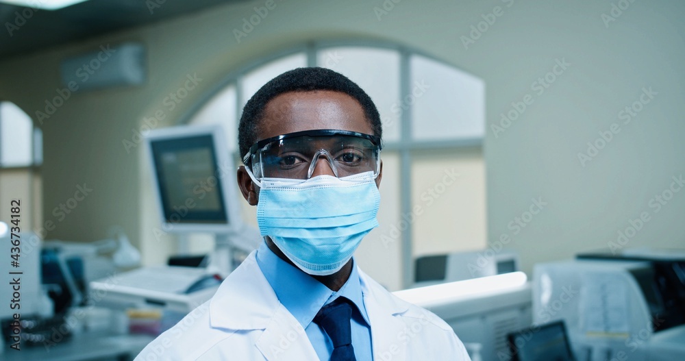 Close up of African American young man healthcare specialist in medical mask looking at camera standing in hospital lab. Male doctor in clinic laboratory during covid quarantine. Job concept