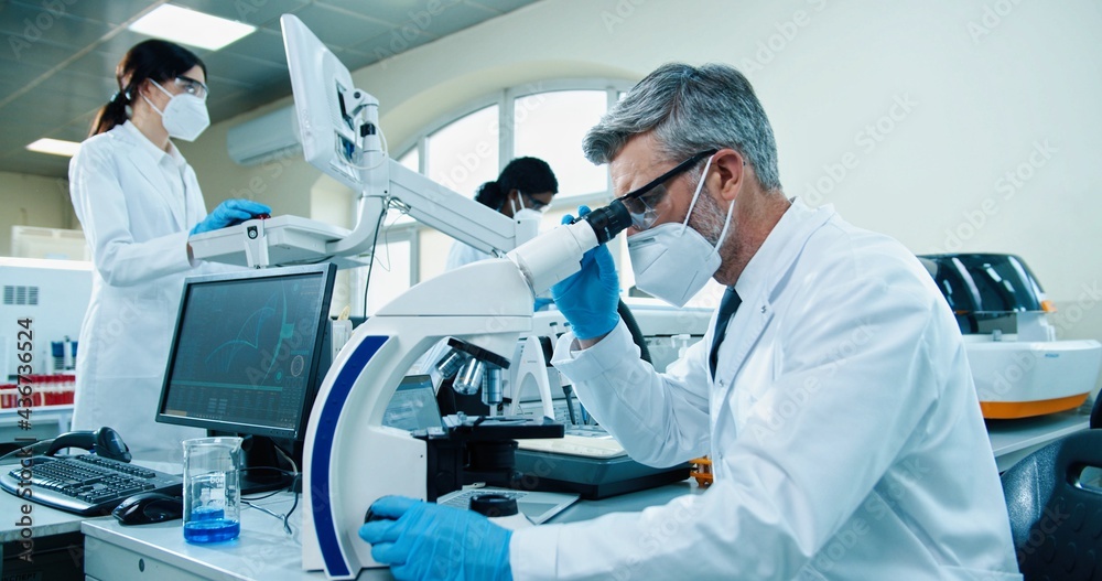Side view of senior Caucasian male medical researcher working in lab on microscope equipment and typing on laptop at workplace, Healthcare worker checking analysis in laboratory, microbiology concept Side view of senior Caucasian male medical researcher working in lab on microscope equipment and typing on laptop at workplace, Healthcare worker checking analysis in laboratory, microbiology concept