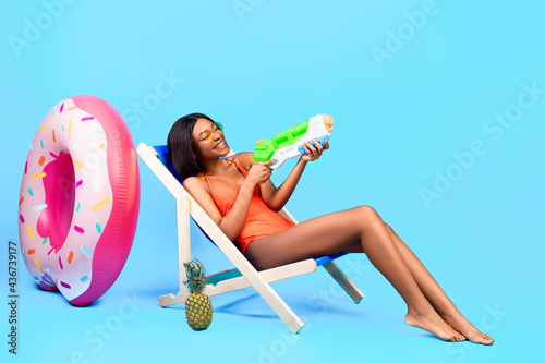 Summertime fun. Attractive black woman sitting in lounge chair with water gun, playing beach game over blue background