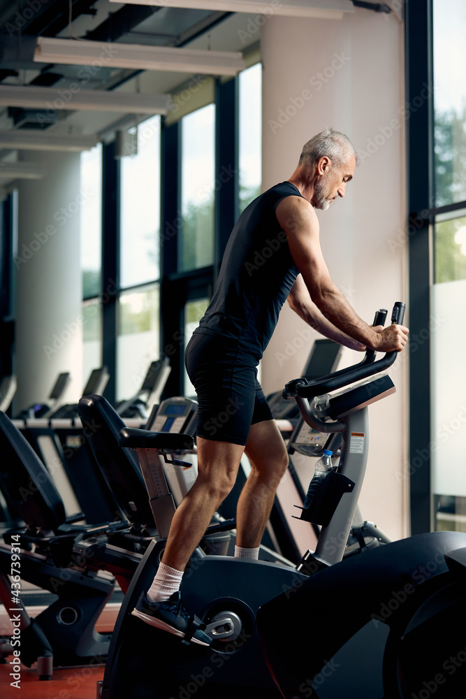 Full length of mature athletic man using exercise bike while working out in a gym. Full length of mature athletic man using exercise bike while working out in a gym.