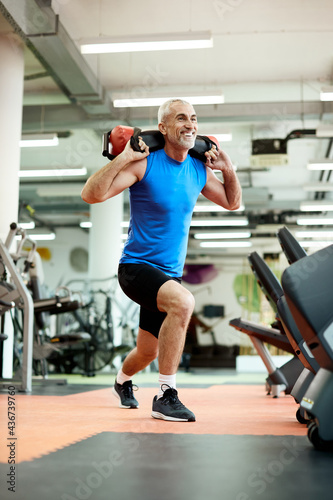 Happy mature sportsman exercising lunges while carrying sandbag on his shoulders in a gym.