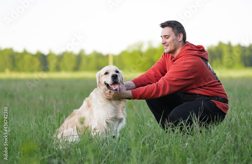 Young man with a golden retriever for a walk in a summer park. Male hands are stroking a joyful dog on the green grass. The concept of interaction with pets.
