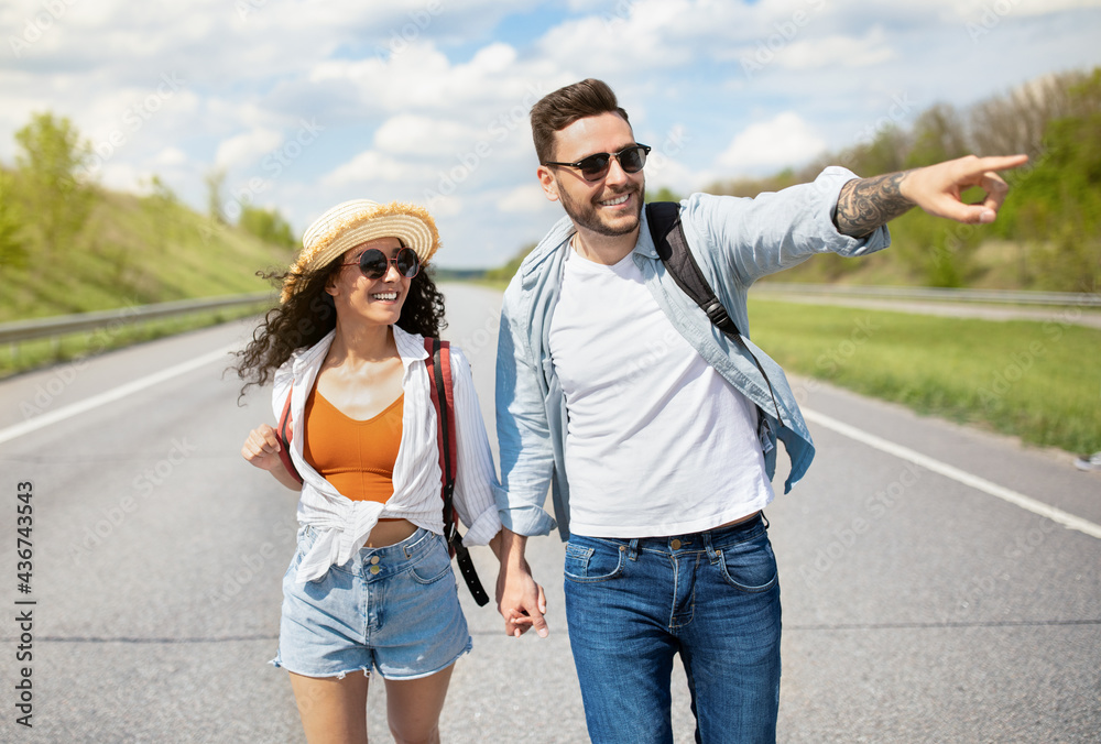 Cheerful young couple with rucksacks walking along road, pointing aside, holding hands, hitchhiking together in summer Cheerful young couple with rucksacks walking along road, pointing aside, holding hands, hitchhiking together in summer
