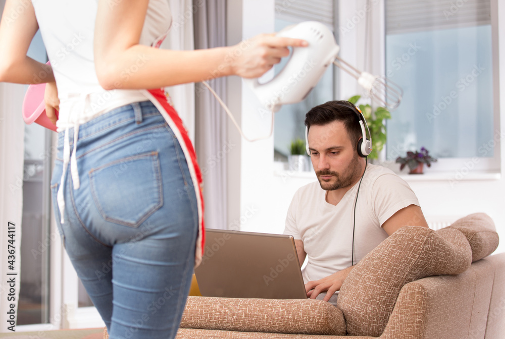 Fototapeta premium Woman doing housework cooking looking at man sitting gaming using laptop