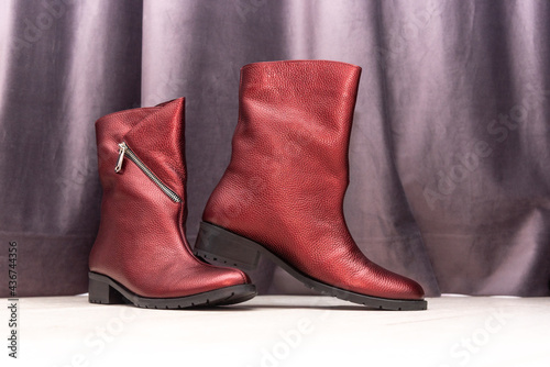Pair of elegant shining red leather women boots with zipper and low heels on white table near purple curtain in light room closeup