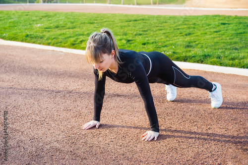 Young fitness woman in black sportswear doing plank. Outdoors workout in the stadium at sunset. Active lifestyle, sport and health care. Copy space
