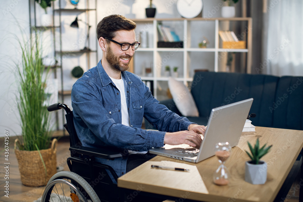 Young man with special needs in casual clothes and eyeglasses working on wireless laptop. Male freelancer working from home while sitting in wheelchair.
