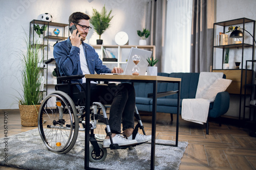 Disabled young man talking on smartphone and working on wireless laptop at home. Male freelancer sitting in wheelchair and using modern gadgets.