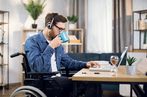 Side view of young man with special need using wireless laptop for remote work at home. Bearded guy sitting in wheelchair  drinking coffee and typing on computer.